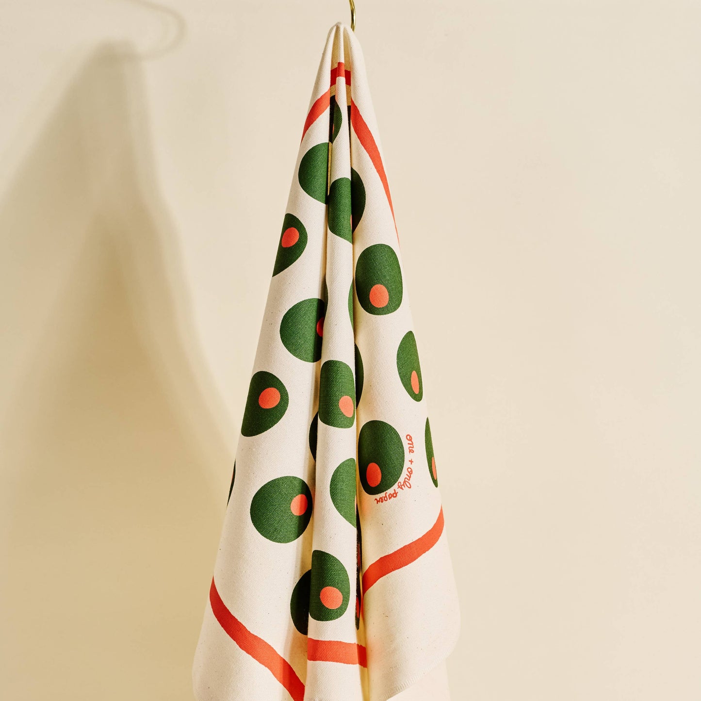 Decorative towel with pimento stuffed green olives pattern, hanging on a hook against a beige wall.