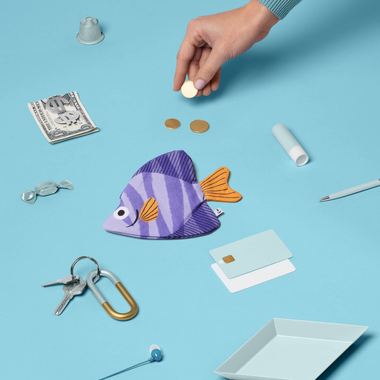 Hand placing a coin into a purple fish-shaped pouch on a blue background with various items around.