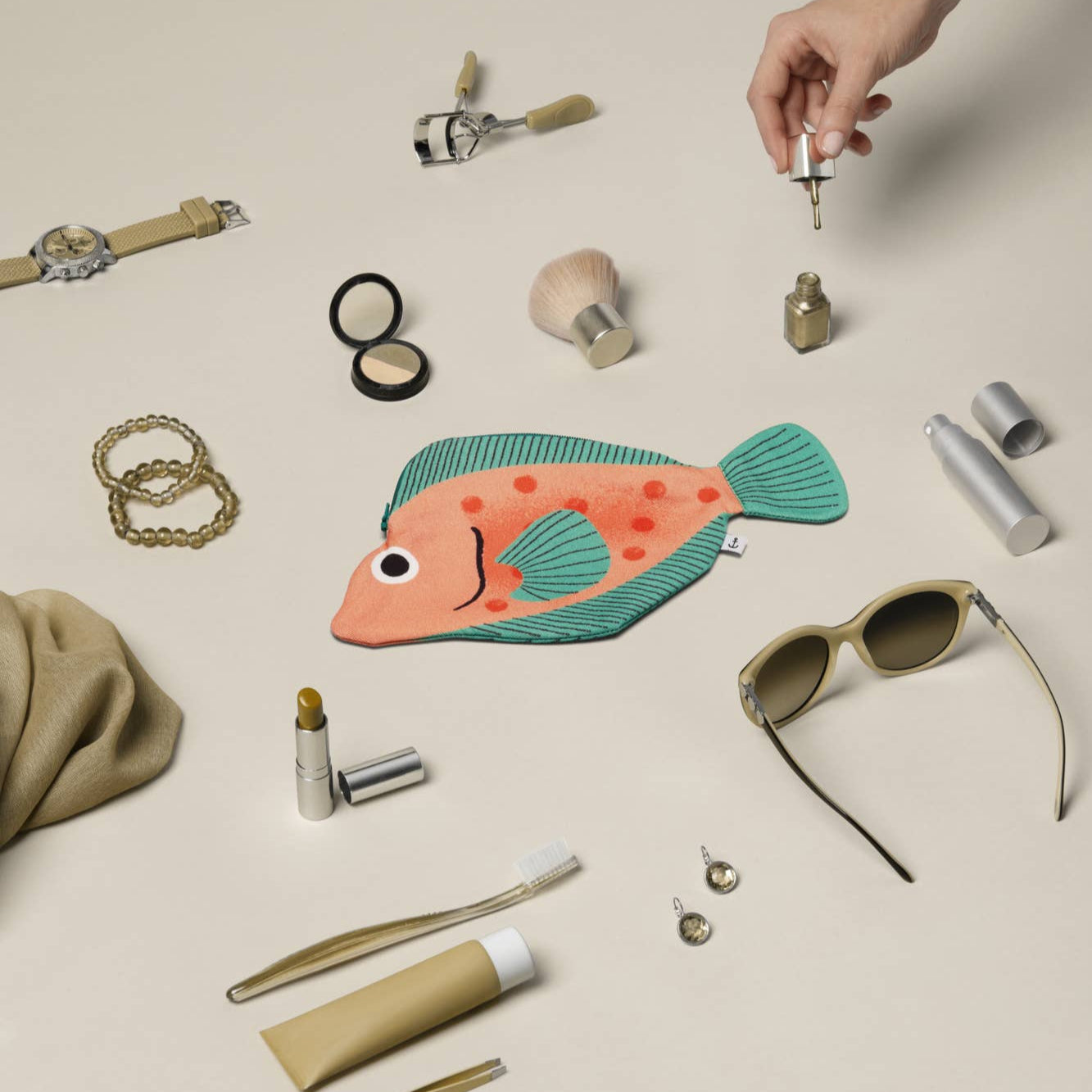 Collection of small items including a fish-shaped pouch, sunglasses, and cosmetic products on a beige surface.