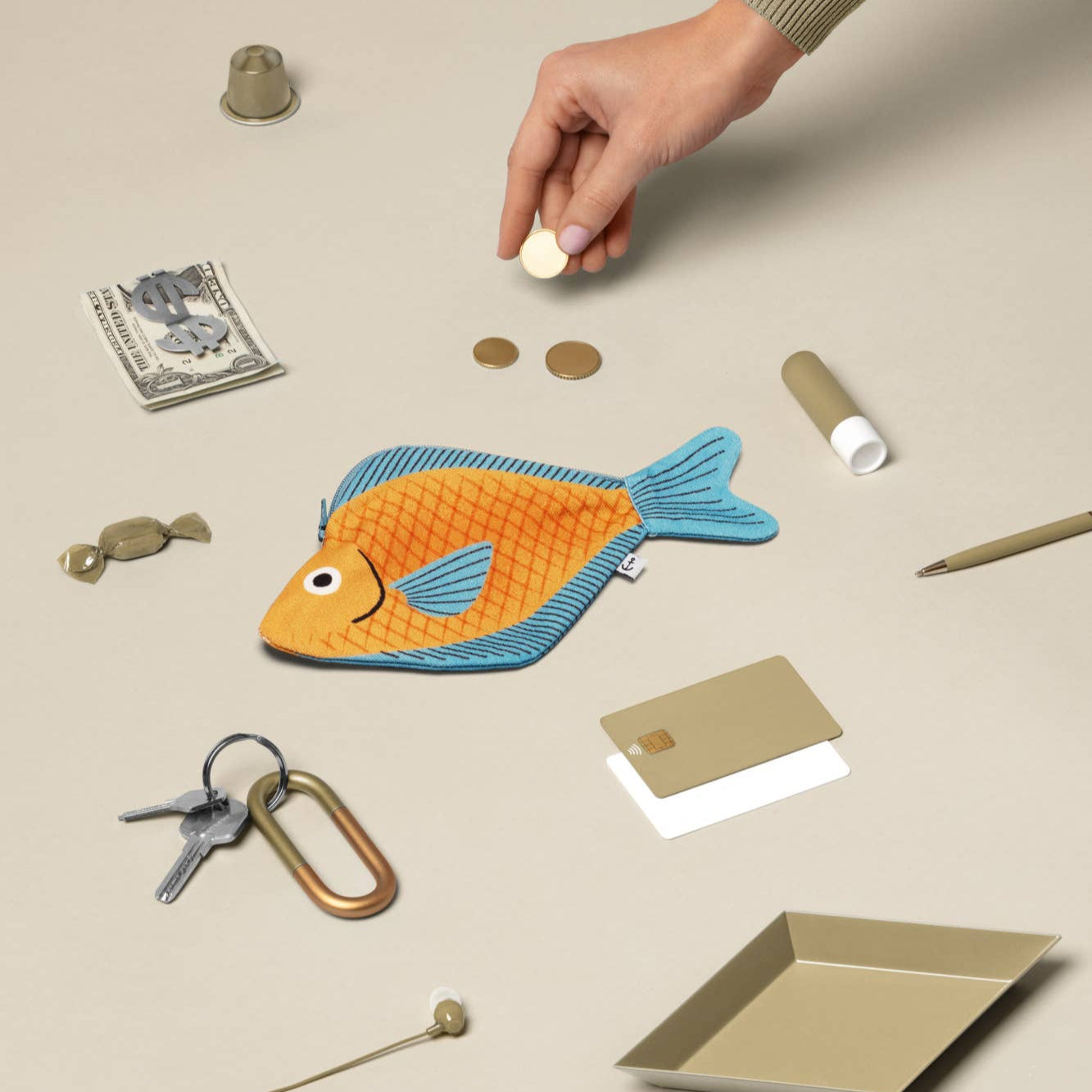 Hand interacting with a fish-shaped pouch on a beige surface with various items around