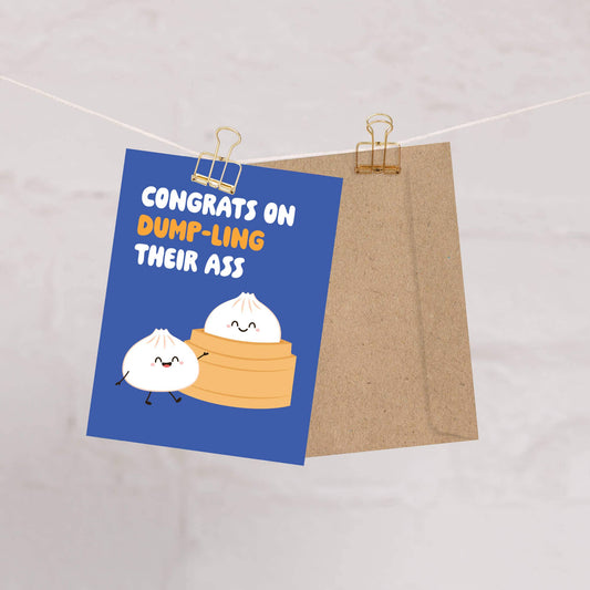 Card with cartoon dumplings and humorous text, "congrats on dump-ling their ass", on a blue background, clipped to a string.