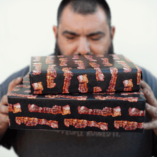 Man wearing a black shirt holding two gift boxes stacked on top of eachother. The gifts are wrapped in bacon patterned gift wrap. 