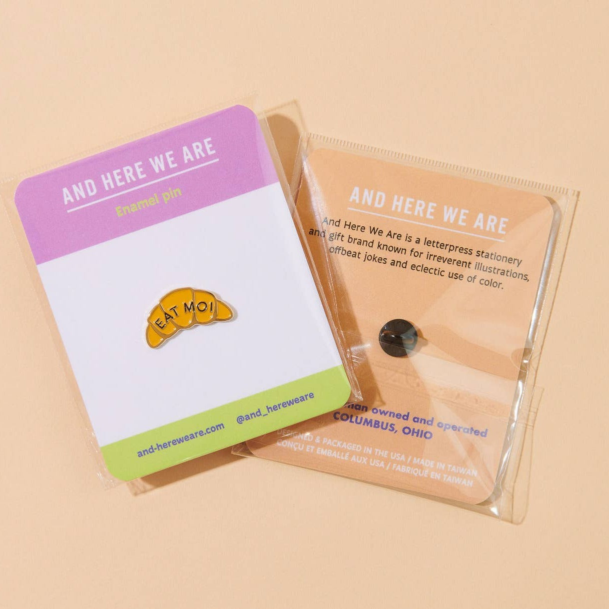 Enamel pin in the shape of a croissant that says "EAT MOI". The lapel pin is staged on a card backing and the picture shows the back and front of the pin with the logo "and here we are"
