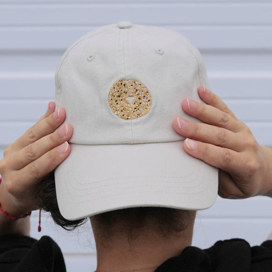 Person holding a beige cap with an everything bagel design against a neutral background