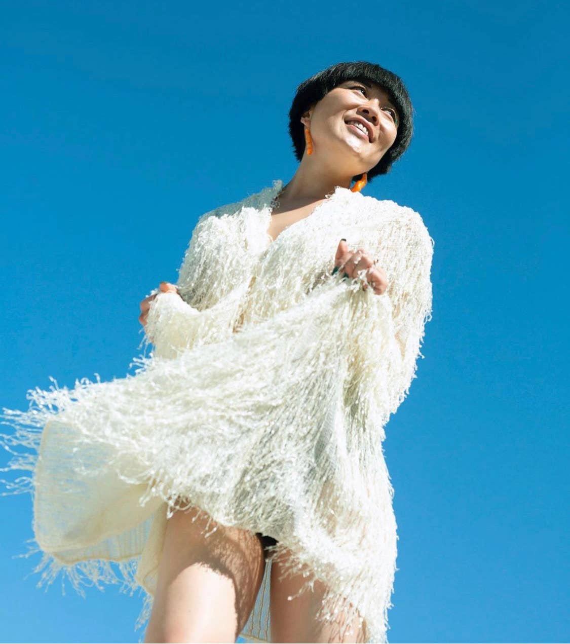 Editorial style photograph of woman wearing the pair of regular cheeto earrings. Woman is wearing a white, fringey dress set against the blue sky