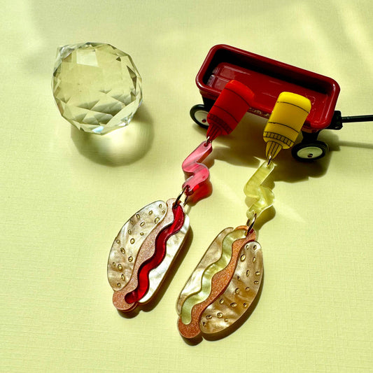 Pair of earrings resembling hot dogs on a light yellow background with a small red toy wagon.