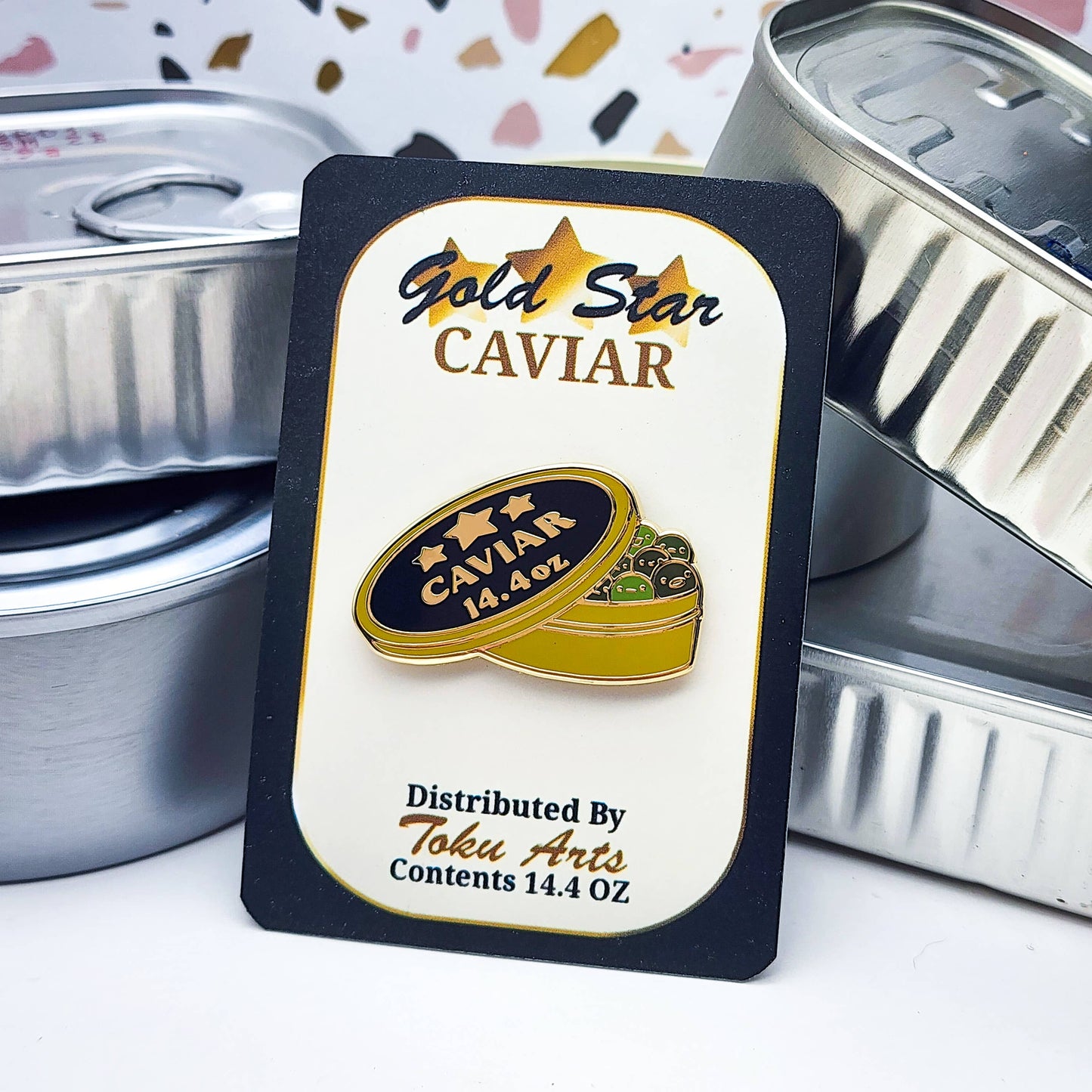 Gold Star Caviar labeled card packaging with the green caviar enamel pin on it, set against plain tinned fish cans in the background