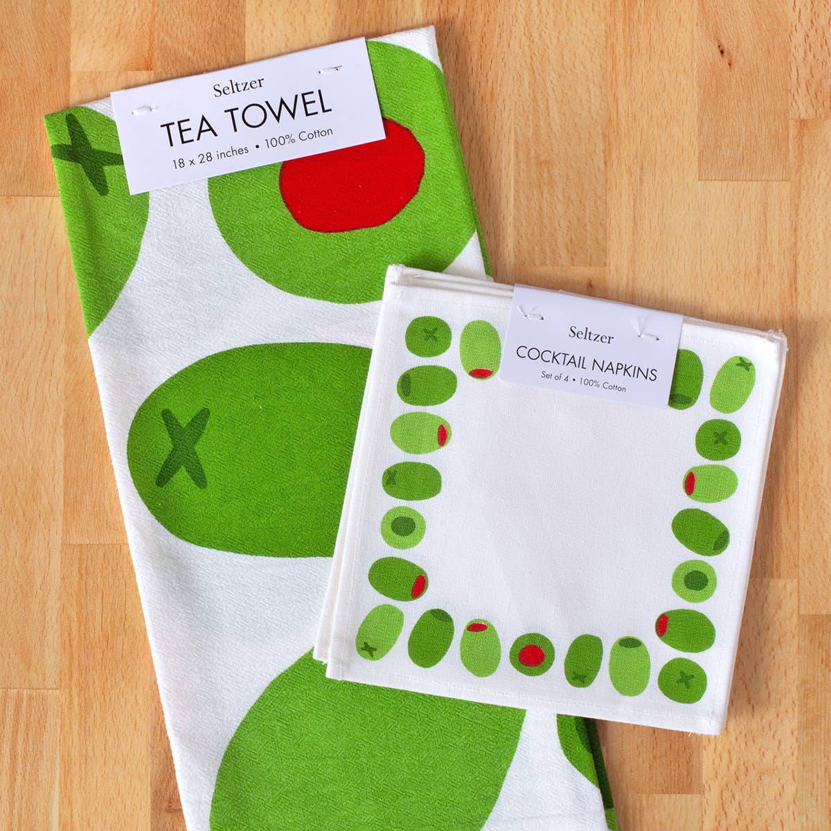 Tea towel and cocktail napkins with stuffed green olive designs on a wooden surface