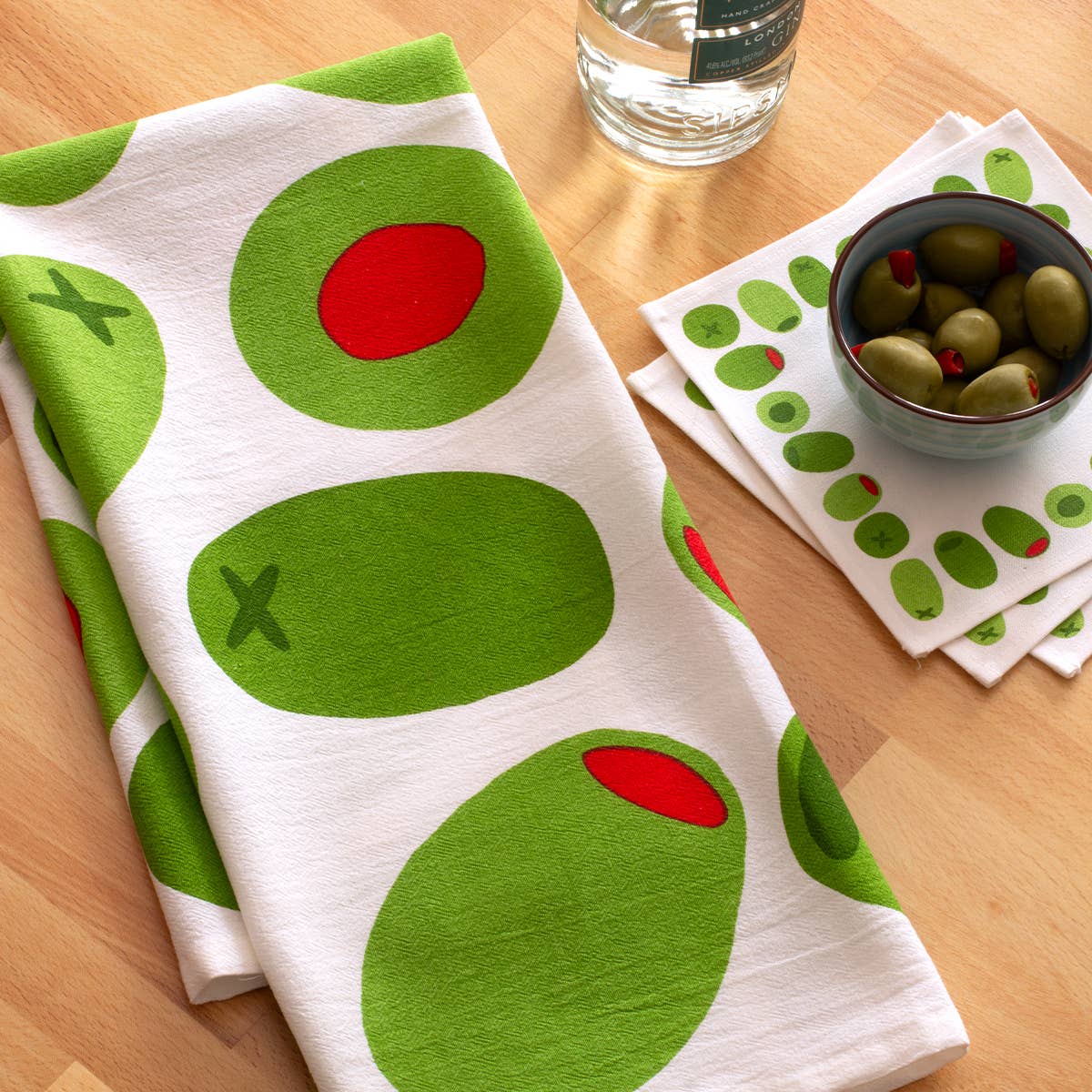 Olive-themed kitchen towel and coasters on a wooden surface with a bowl of olives.