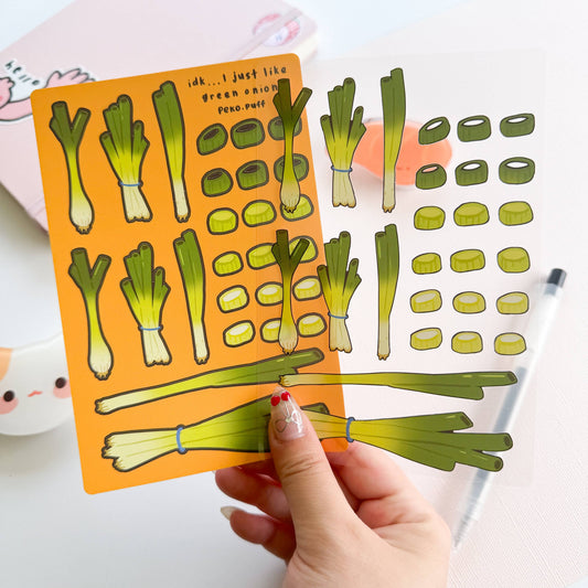 Hand holding a sticker sheet with green onion illustrations on an orange background