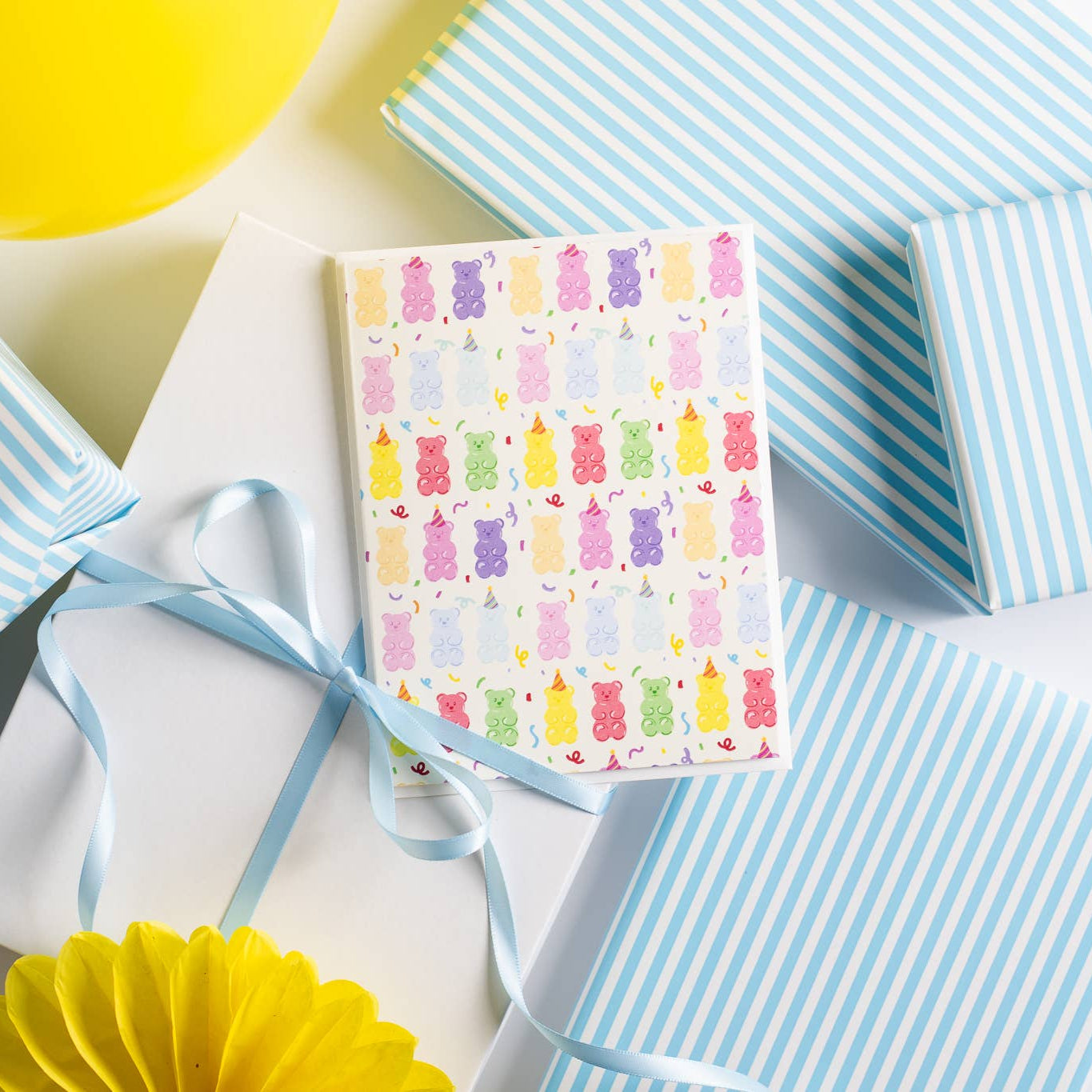 Greeting card with colorful gummy bear pattern surrounded by blue and white striped gift boxes on a white background.