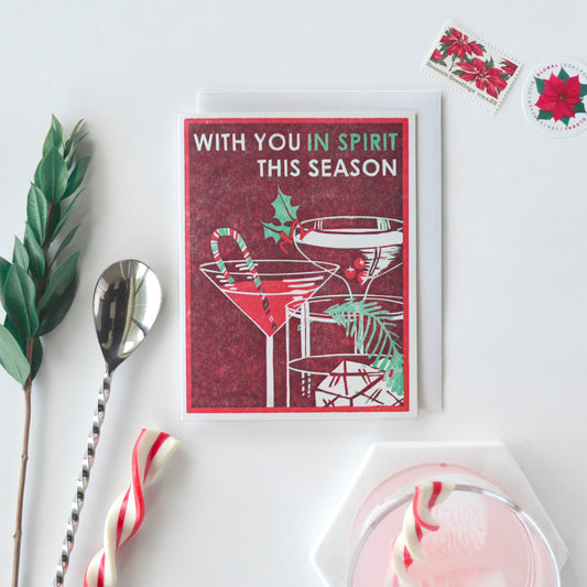 holiday card that reads "with you in spirit this season" photographed with other various objects including, holiday stamps, a peppermint twist, coaster, cocktail spoon