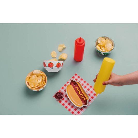 Hot dog shaped plate with mustard, ketchup, and chips on a checkered napkin with a hand holding a yellow mustard bottle.