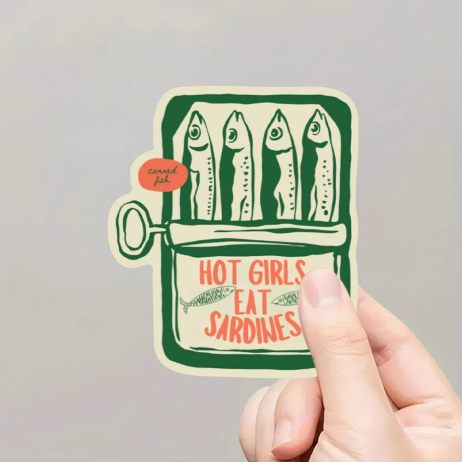 Sticker with sardines design and text held by a hand against a gray background