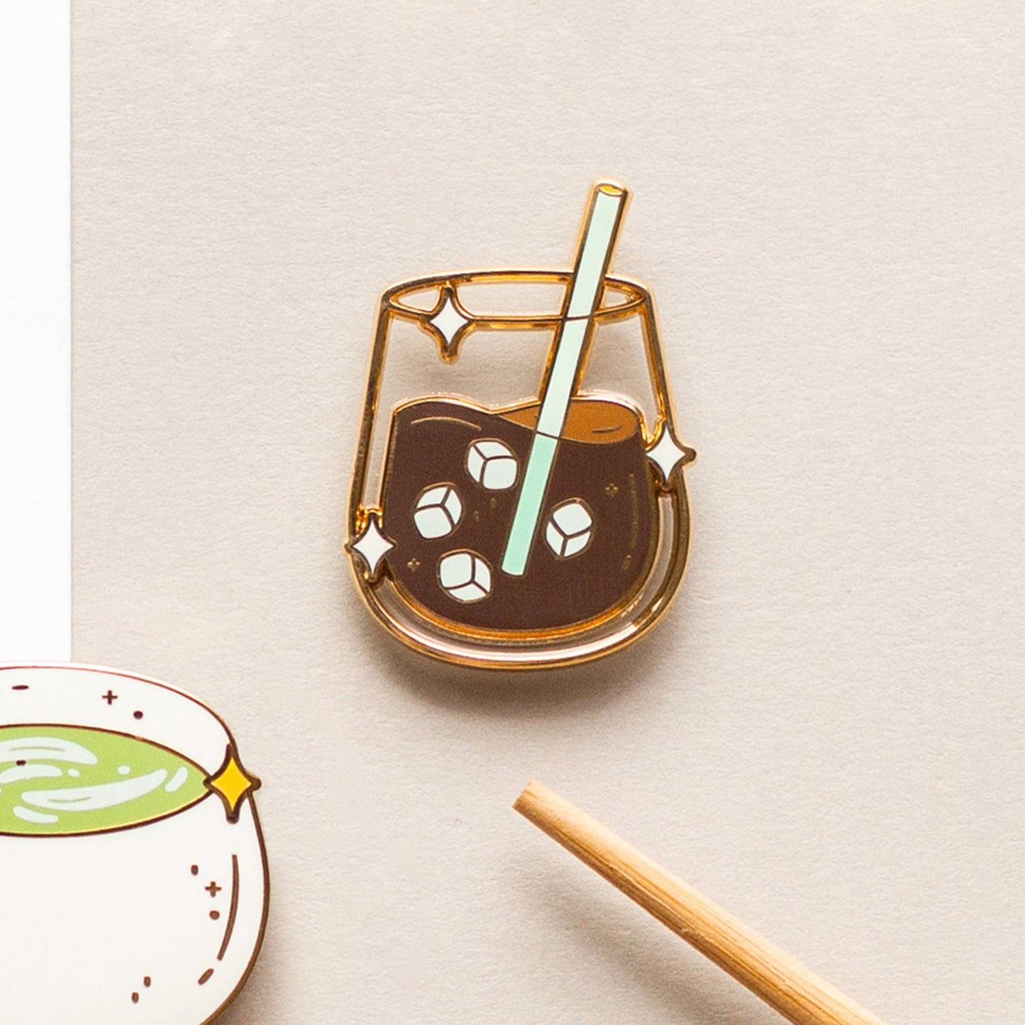 Enamel pin of a glass of iced coffee with a straw on a beige background