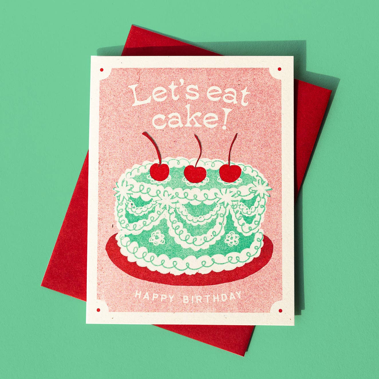 Birthday card with green frilly cake, decorated with ribboned icing and three red cherries on the top. Text reads "Let's eat cake! Happy birthday"