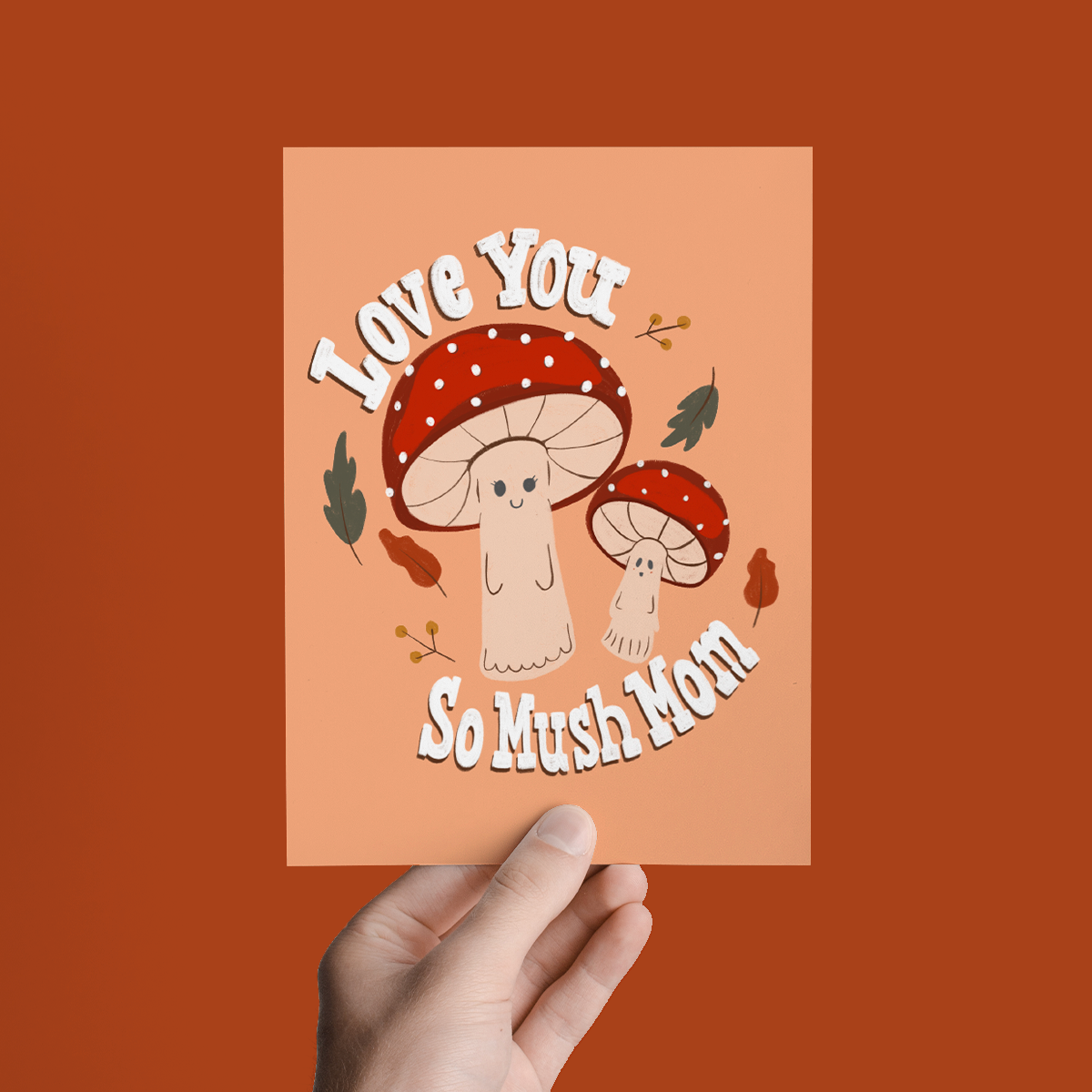 Card with cartoon mushrooms and text 'Love You So Mush Mom' held against an orange background
