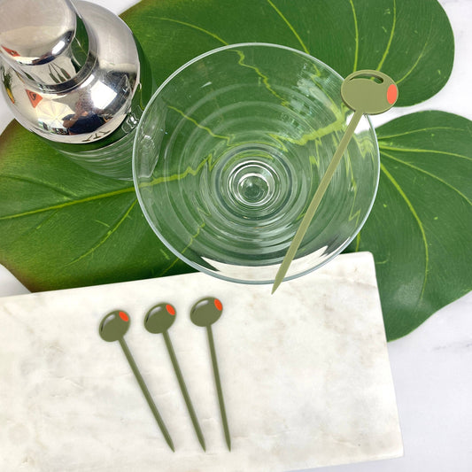 Clear glass with olive cocktail stirrers on a white napkin and green leaves