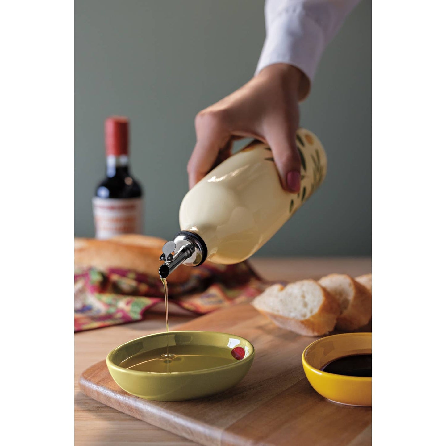 Person pouring olive oil from a bottle into a green bowl on a wooden table with bread and a bottle in the background.