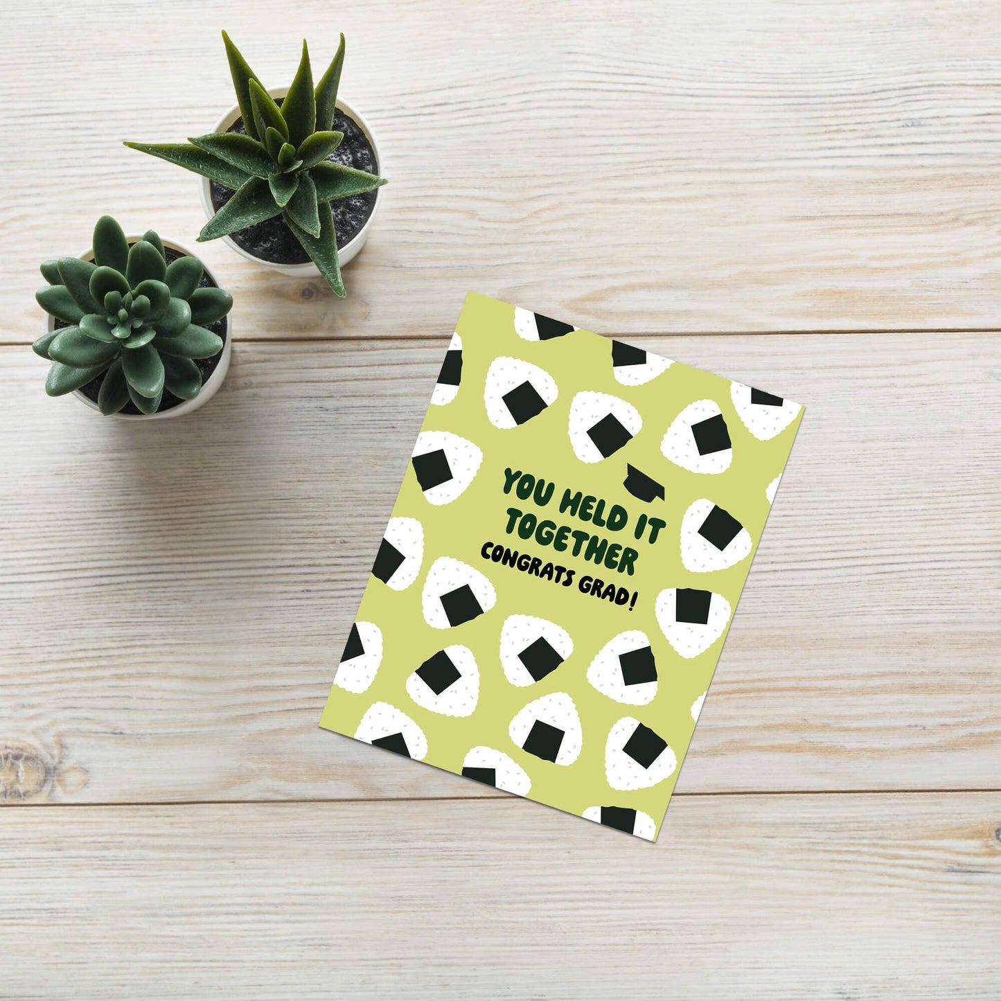 Graduation card with onigiri rice ball pattern on a wooden surface with small plants.