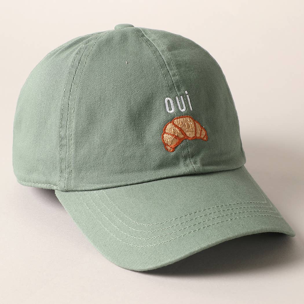 A green baseball cap with 'oui' written on it and an embroidered croissant below the text.