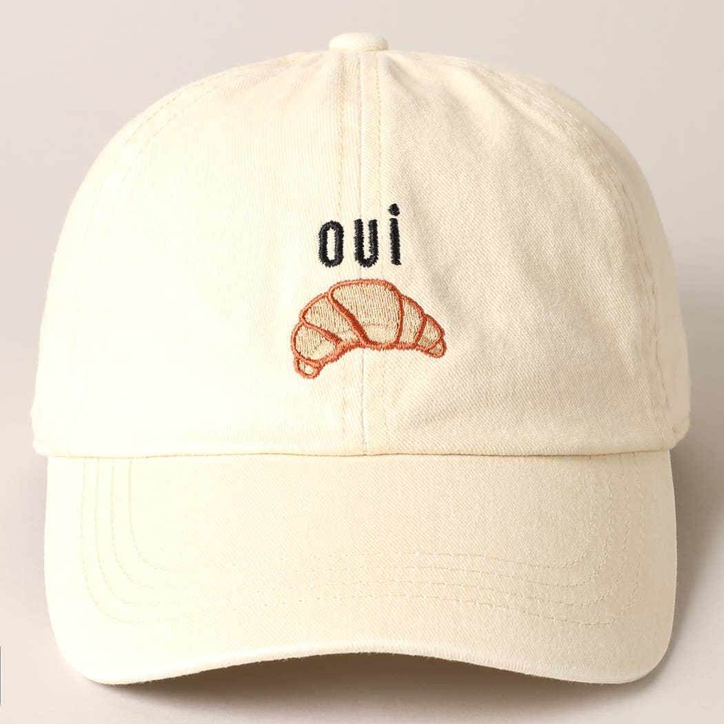 A cream baseball cap with 'oui' written on it and an embroidered croissant below the text.