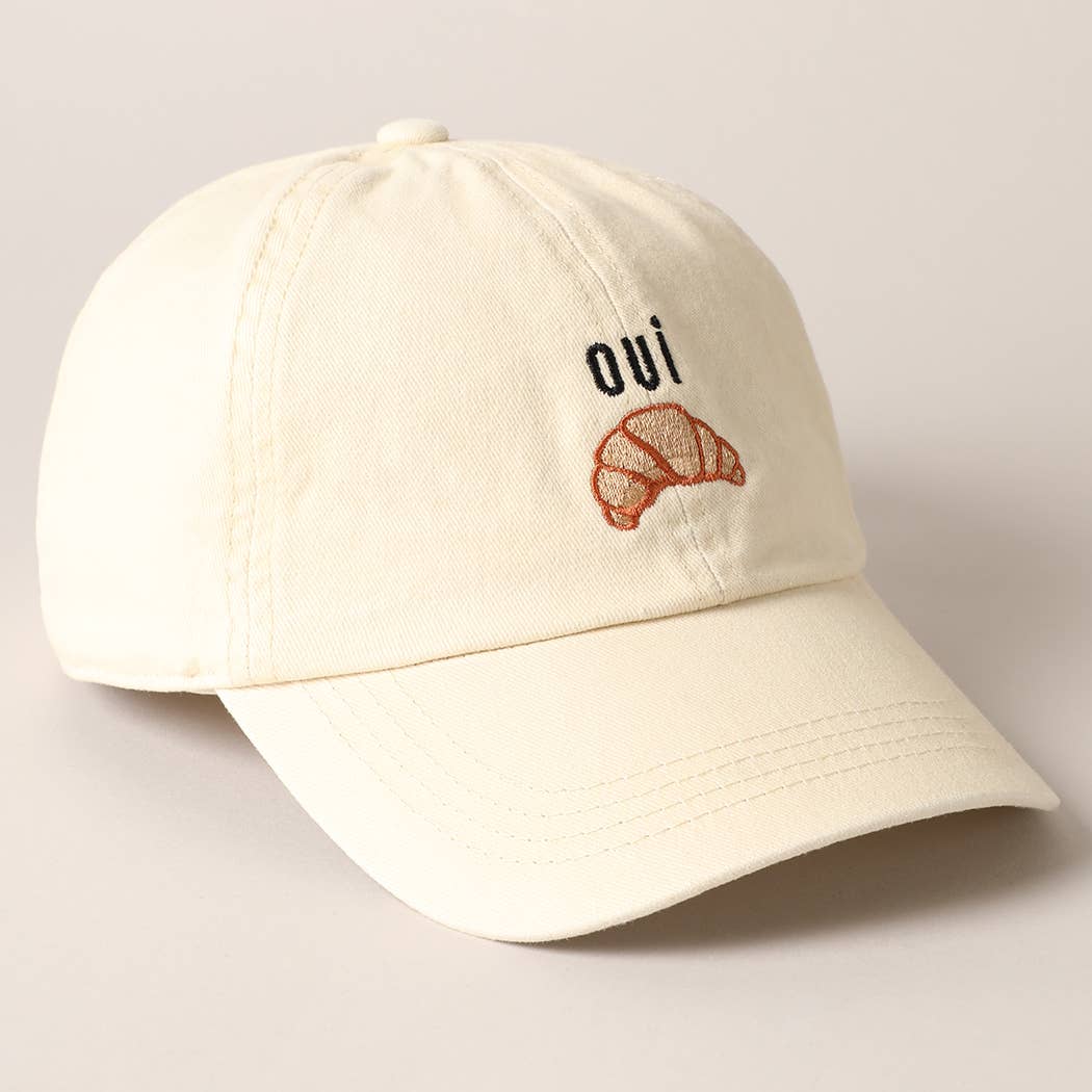 A cream baseball cap with 'oui' written on it and an embroidered croissant below the text.