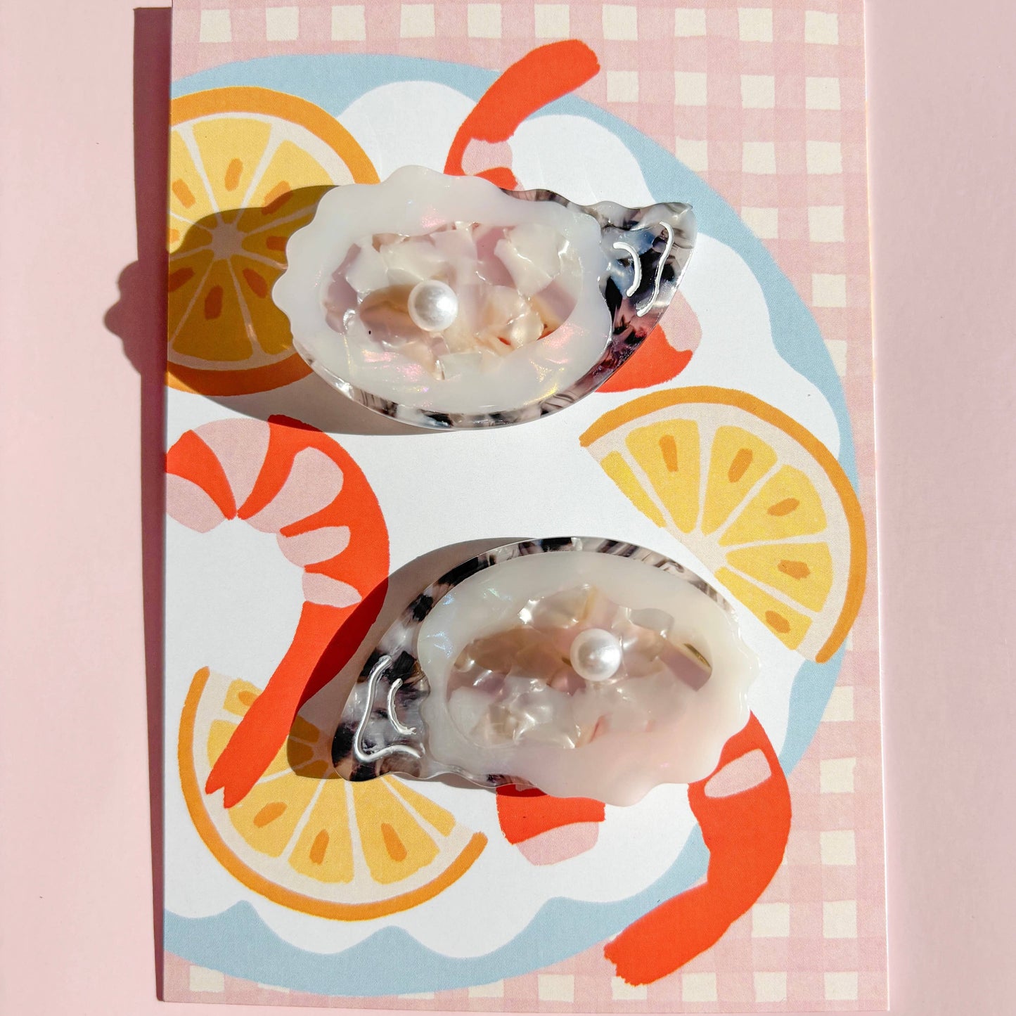 Pair of oyster shaped hair clips on a decorative patterned paper with shrimp and lemon design on a pink background