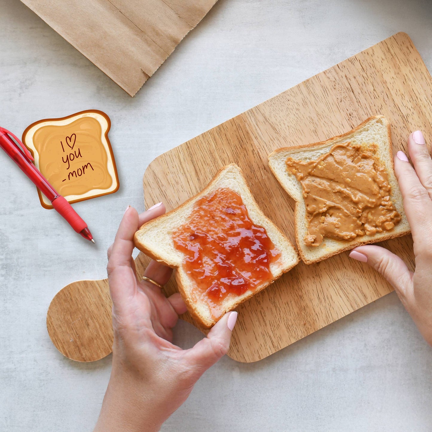Two slices of toast with peanut butter and jelly on a wooden cutting board, with hands arranging the toast and a peanut butter sticky note in top left corner that says "I <3 you -Mom"