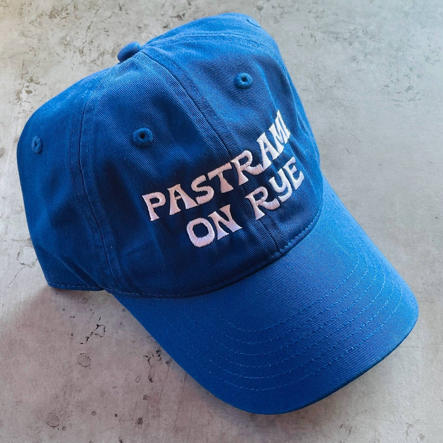 blue baseball cap with white embroidered text that reads "pastrami on rye"
