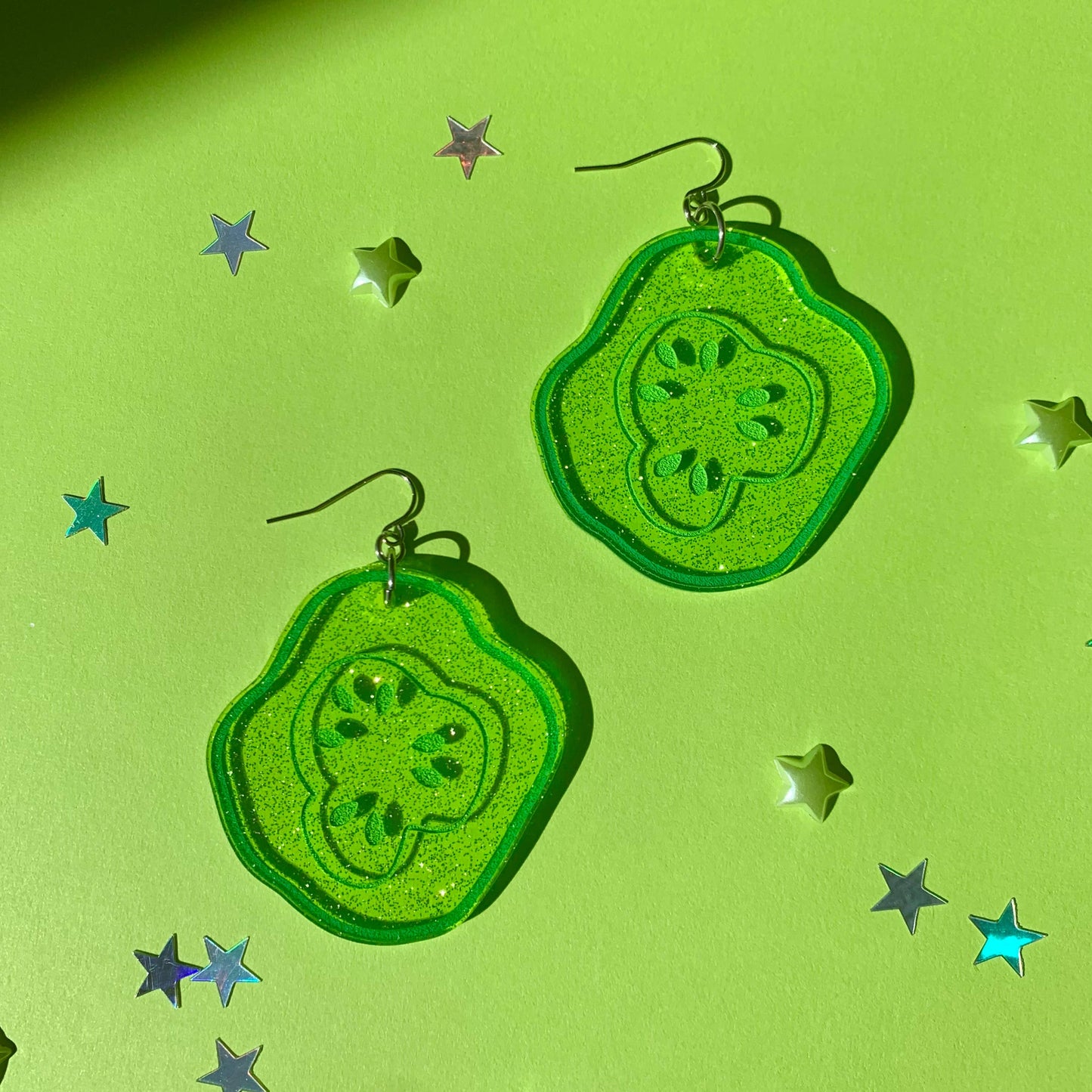 Glittery, green, acrylic, sliced pickle earrings on fish hook backings on green background with mini star confetti pieces