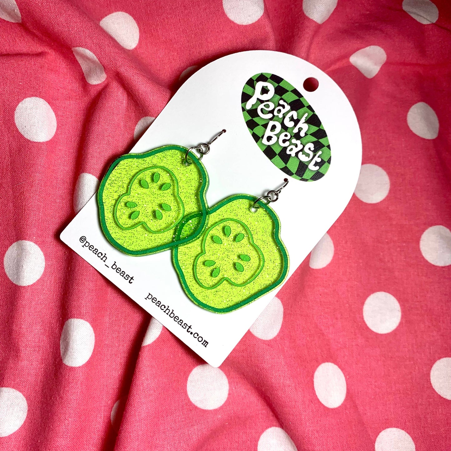 Glittery, green, acrylic, sliced pickle earrings on fish hook backings with cardboard backing/packaging. Photographed on pink and white polka dot background
