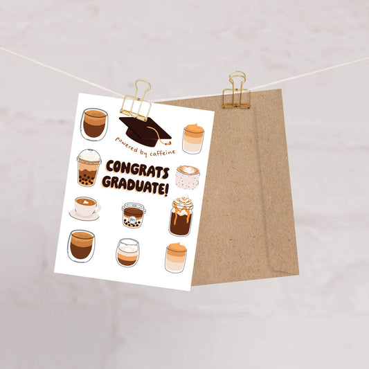 Graduation card with coffee illustrations and 'Congrats Graduate!' text, attached to a brown envelope with gold clips.