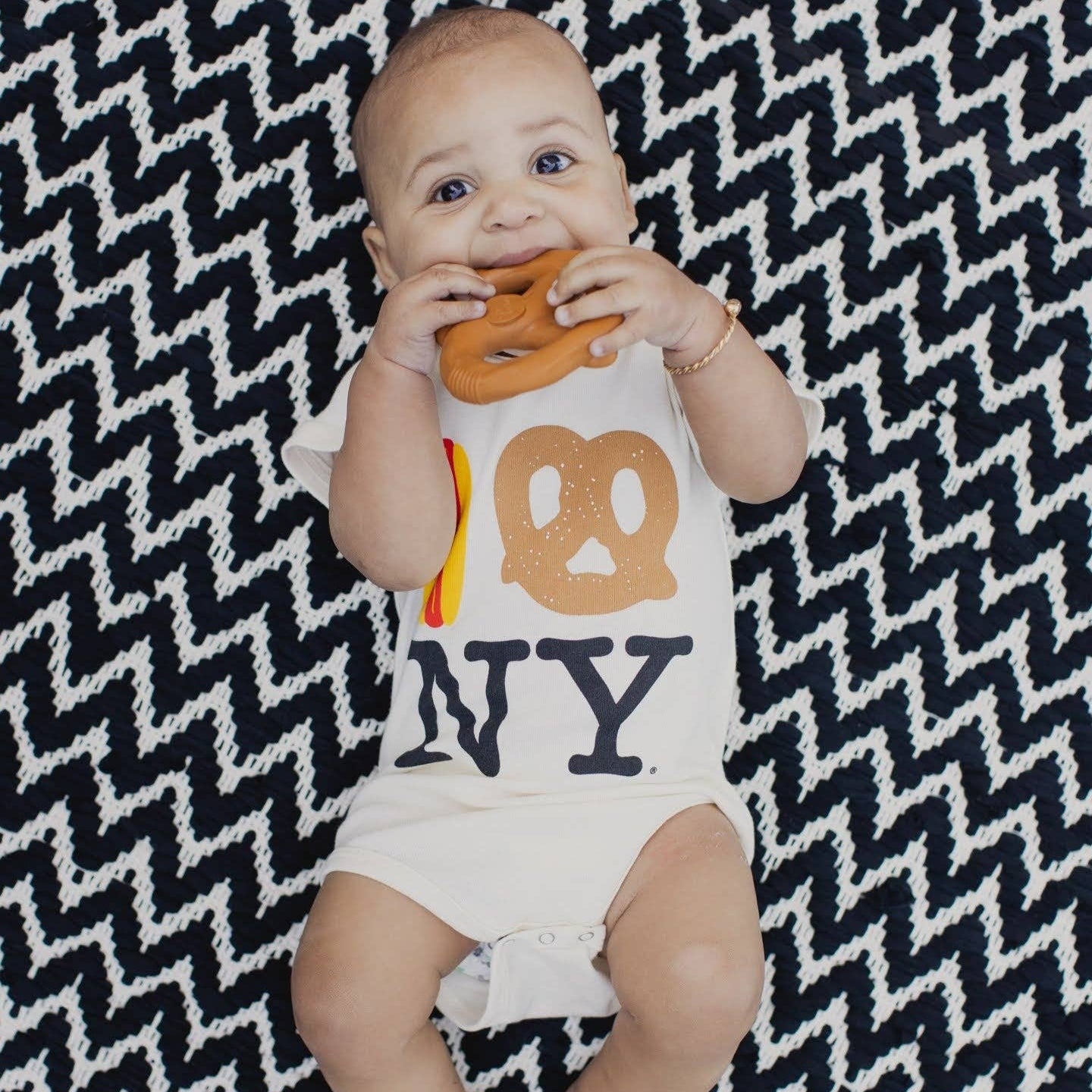 Baby wearing a onesie with 'NY' and pretzel design, sitting on a black and white zigzag patterned blanket with the pretzel teether in it's mouth.
