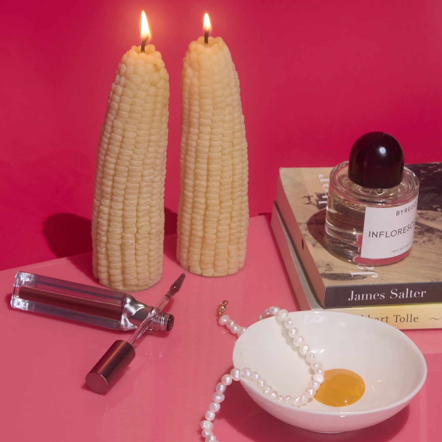 Two lit corn-shaped candles on a pink surface with the egg bowl, perfume, lip gloss, necklace, and books.