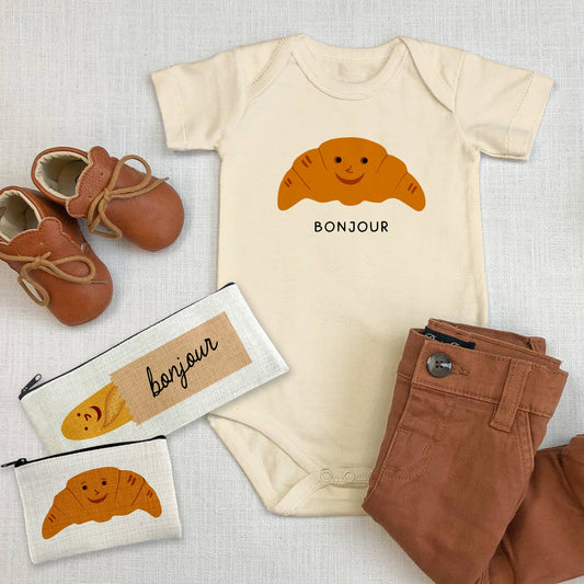 flat lay of the bonjour croissant baby onesie, brown baby pants and shoes and two small pouches one with a baguette and the other with the same smiley croissant on it