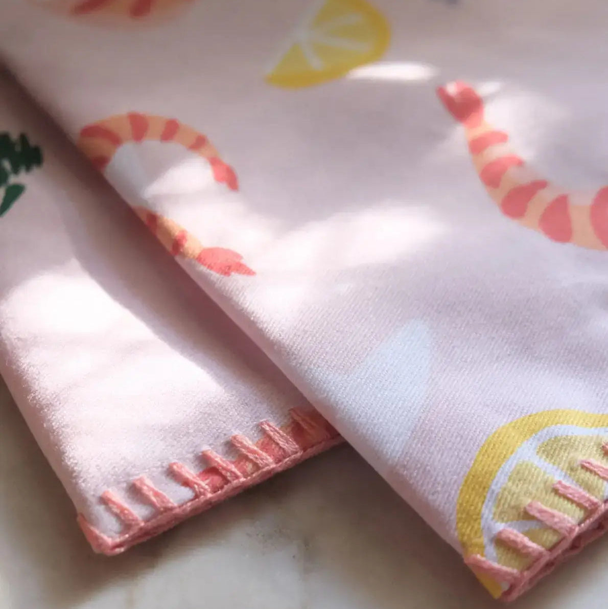close up of the kitchen tea towel, light pink/salmon in color with a design of illustrated lemon halves and wedges, shrimp, cocktail fork and herbs