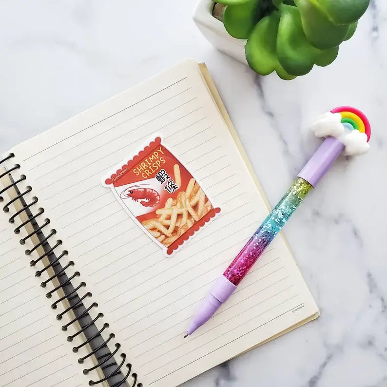 Sticker made to look like a bag of Asian shrimp chips, laying on top of a lined notebook with a rainbow pen