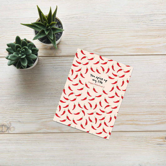 Greeting card with red chili pepper pattern and text "you spice up my life. Happy Valentine's!" on a wooden surface with small plants