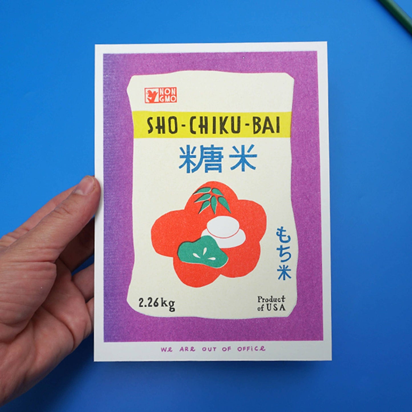 Hand holding a risograph art print with a Shokubai product label illustration against a blue background
