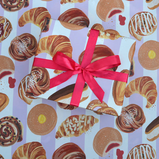 sheet of gift wrap with a striped, lilac and white background and different sweet bakery treats on it including croissants, doughnuts and cinnamon rolls
