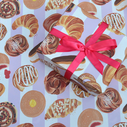 sheet of gift wrap with a striped, lilac and white background and different sweet bakery treats on it including croissants, doughnuts and cinnamon rolls