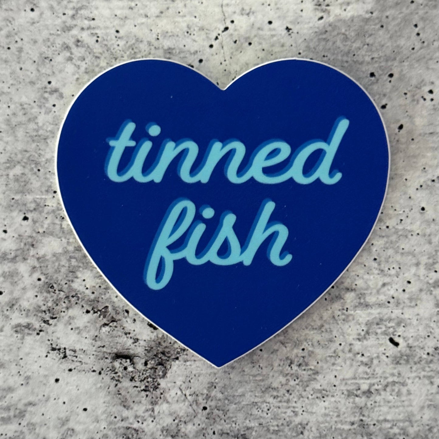 Blue heart-shaped sticker with 'tinned fish' text on a concrete surface