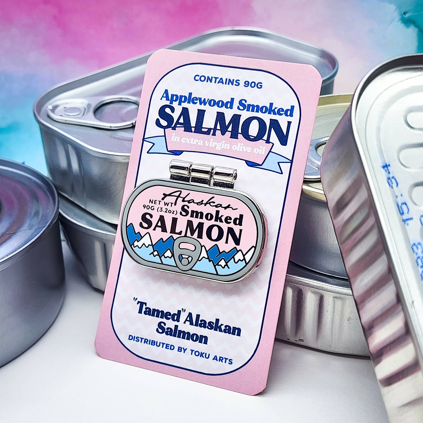 Alaskan Smoked Salmon container as an enamel pin, with mountain design. Pictured here with its card backing, set against tinned fish containers