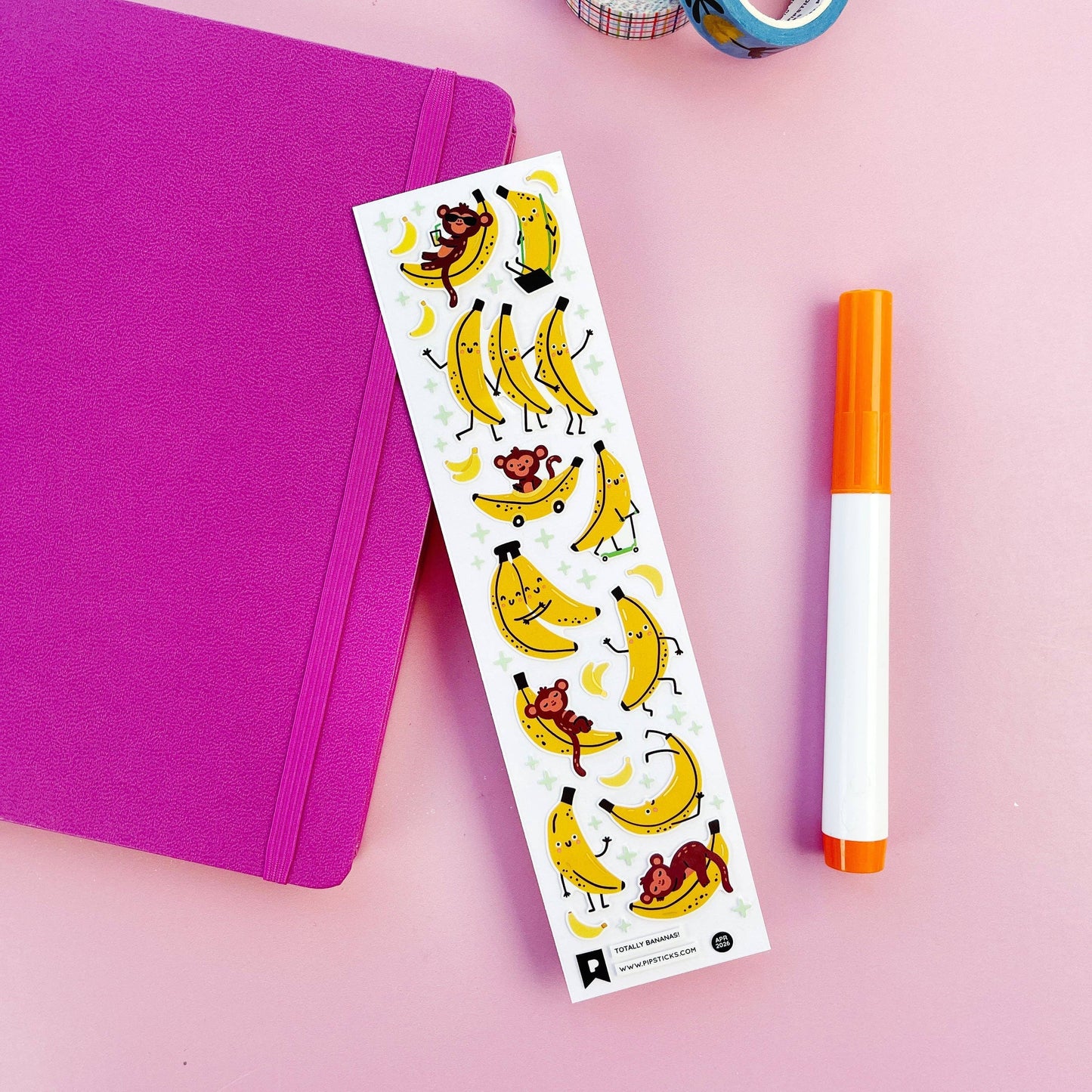 Sheet of stickers with cartoon bananas and monkes on a pink surface with a purple notebook and orange marker.