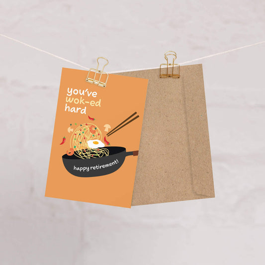 Greeting card with wok and noodles illustration and text that reads "you've wok-ed hard. Happy retirement!" on an orange background, hanging on a string.