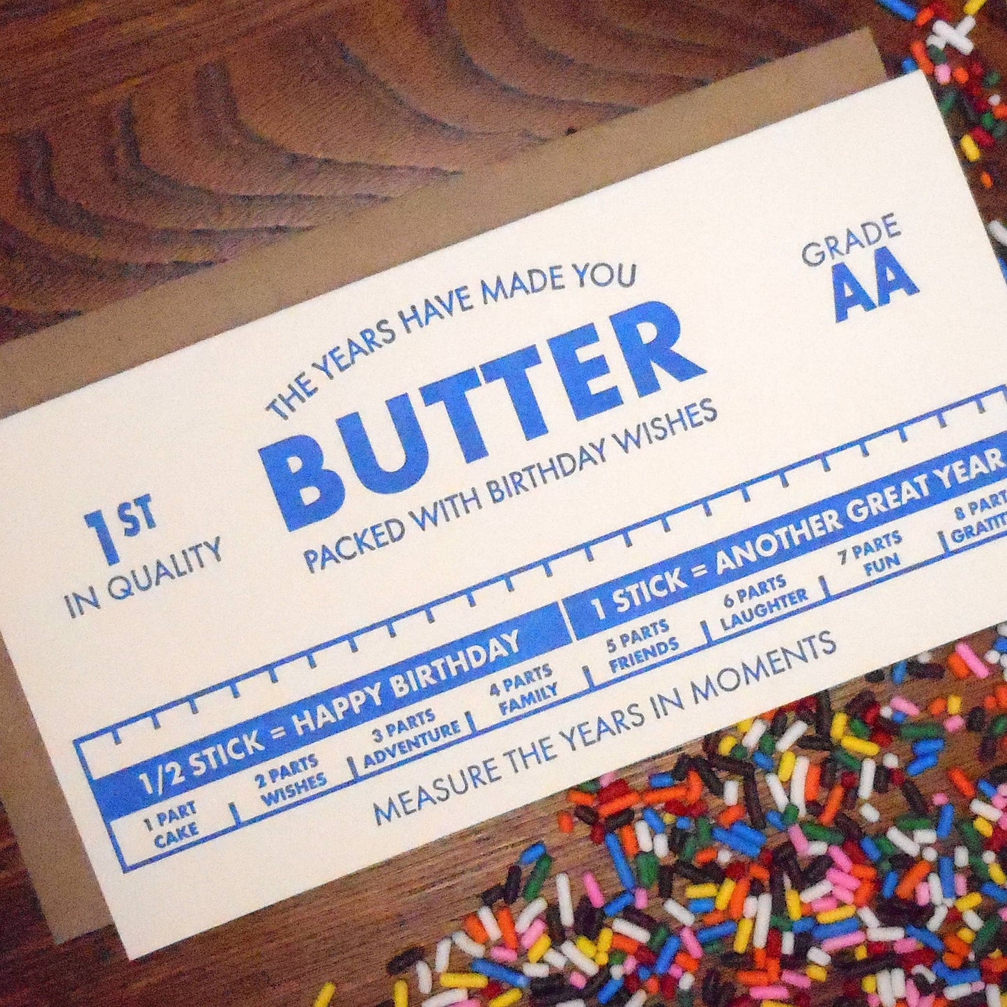 Birthday card with butter text and a ruler graphic on a wooden surface with sprinkles.