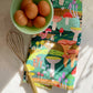 Tea towel with a colorful mushroom design all over it. Shown here with a whisk and bowl of eggs 