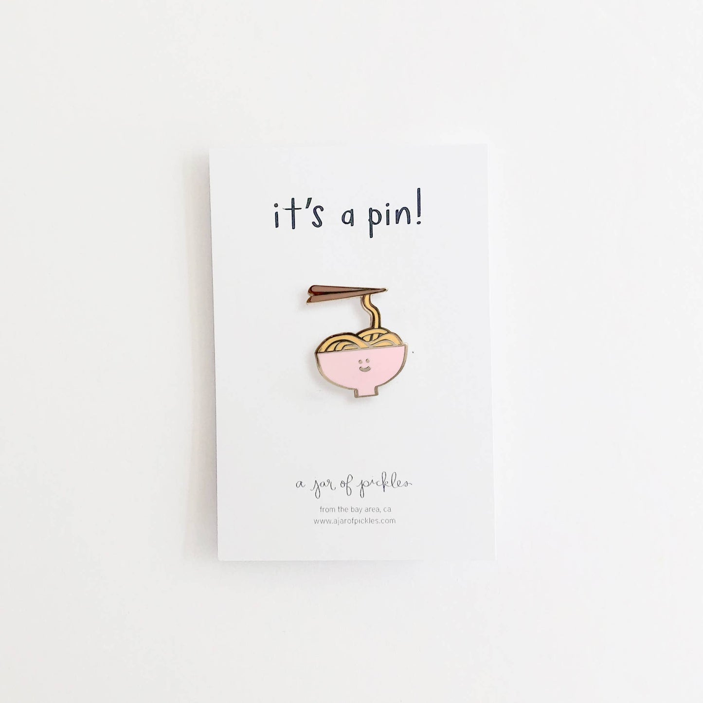 Noodle bowl enamel pin -- bowl is pink with yellow noodles inside, chopsticks are picking up one noodle