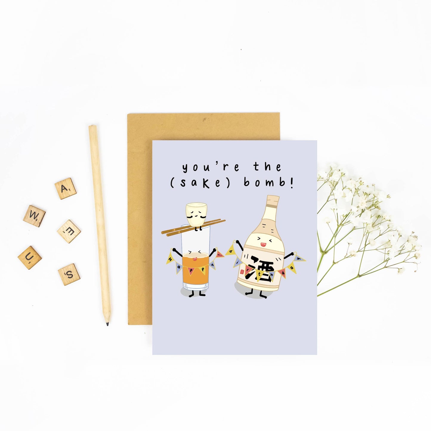 Pale blue greeting card with illustration of a happy anthropomorphic Sake Bottle holding a colorful banner that reads "happy birthday," while a Sake Cup sits precariously on top of chopsticks perched over a glass of beer. Text reads "you're the (sake) bomb!"